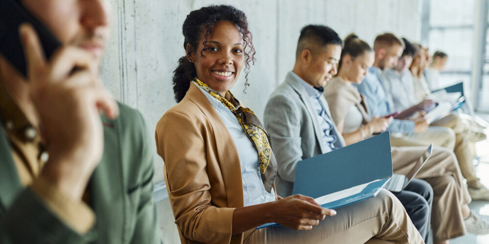 Group of happy, professional people sitting in chairs in a line against a wall.