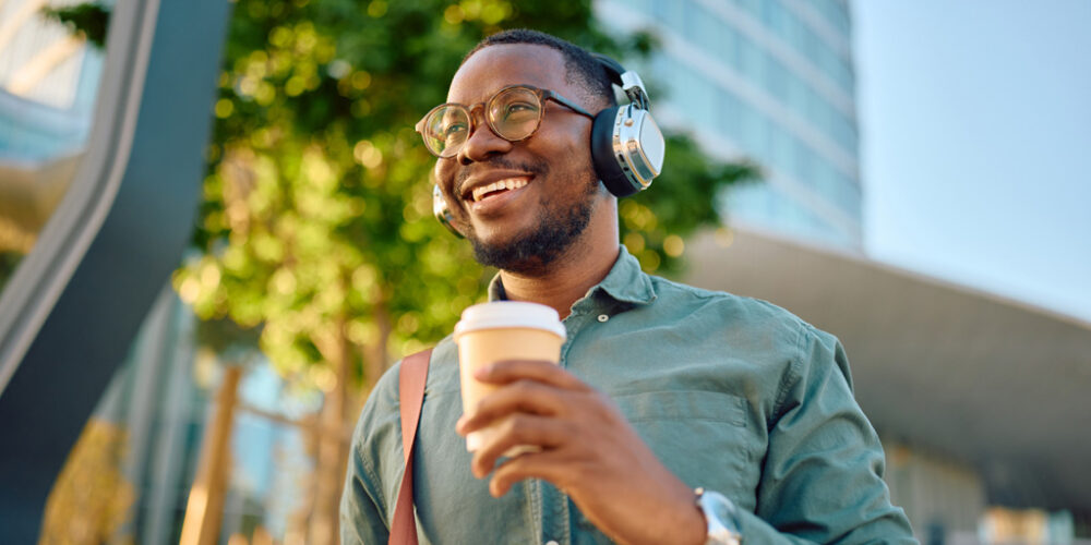 Smiling man wearing headphones and holding a to-go-cup outside.